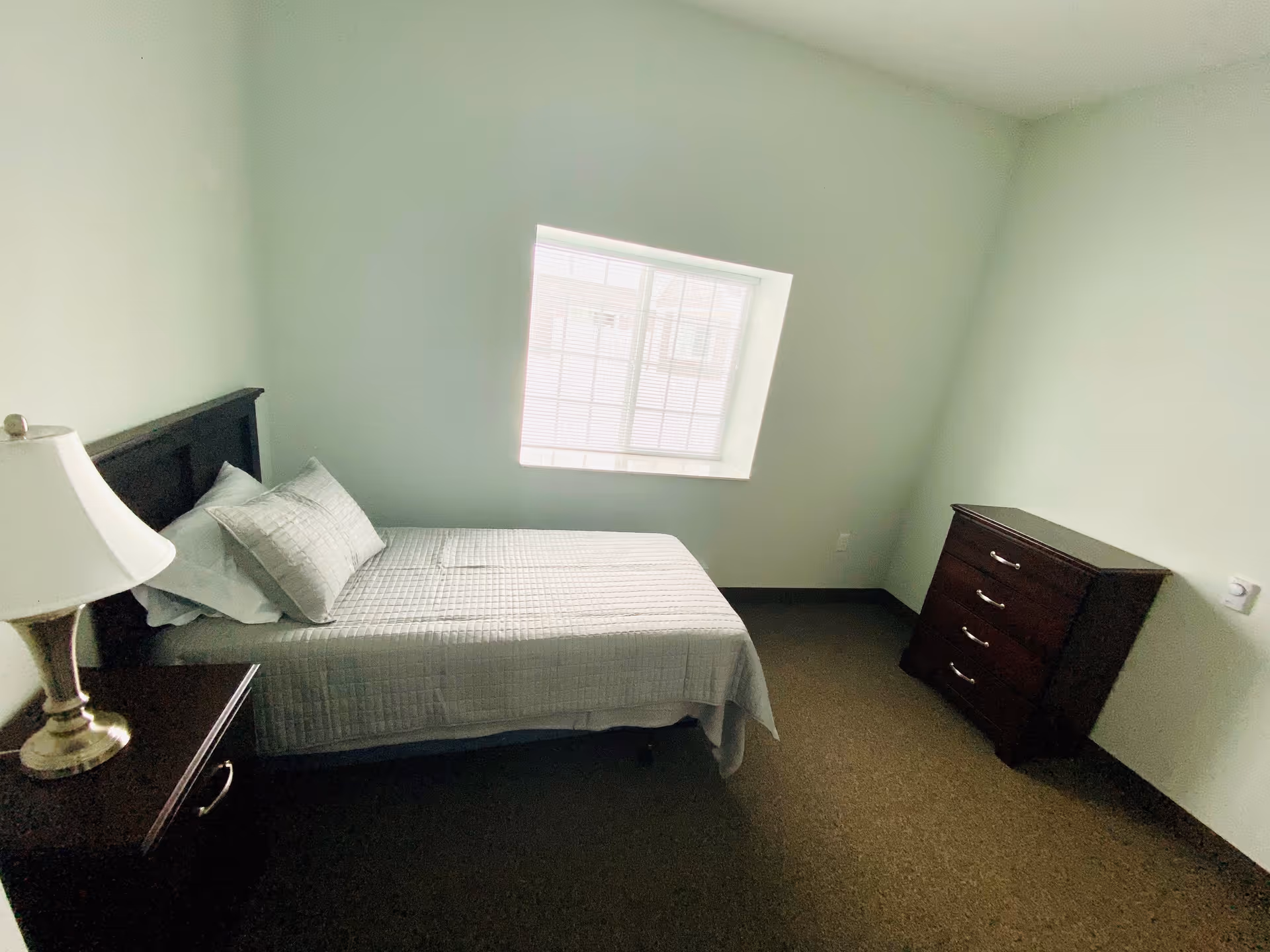 Simple bedroom with a single bed, bedside table and lamp, a dresser, and a window.