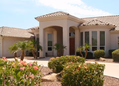 Exterior front view of a single-story building with a tiled roof, arched entrance, and large windows. The landscaping includes small palm trees, bushes, and flowering plants under a clear blue sky.