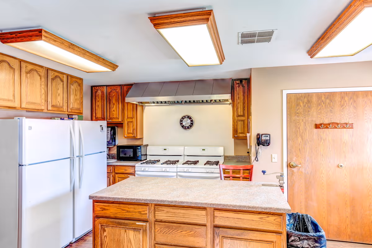 A kitchen with wooden cabinets, a white double-door refrigerator, a microwave, a white stove with a range hood, a wall clock above the stove, a countertop island in the center, a wooden door with hooks, a wall-mounted telephone, and ceiling lights with wooden frames.
