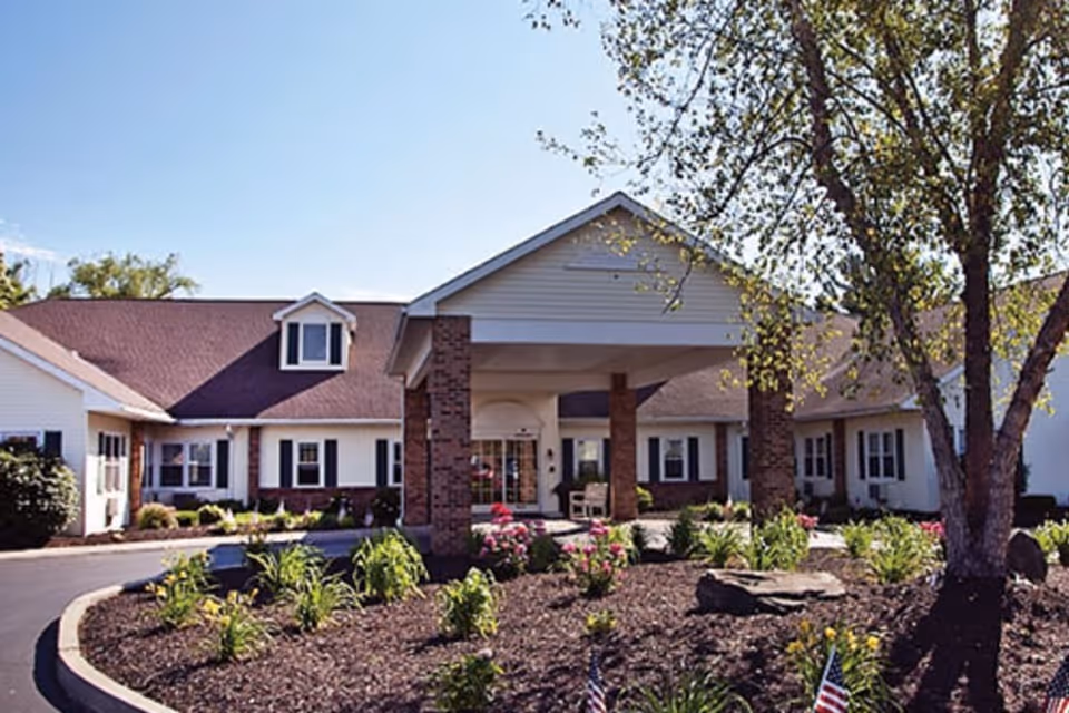 Front exterior of a single-story senior living facility with a covered entrance, brick columns, and landscaped garden.