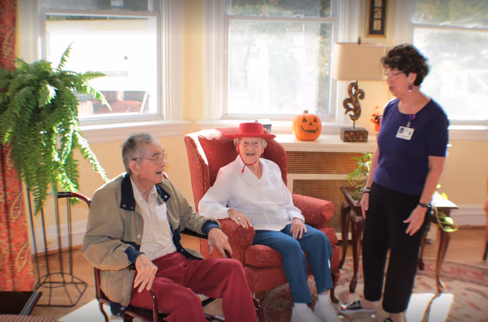 Two elderly individuals sitting in a sunlit room with large windows. One man is seated in a wheelchair wearing a beige jacket and red pants, and a woman wearing a red hat and white blouse is sitting in a red armchair. A woman standing nearby is engaging with them, wearing a navy blue top and black pants with a name badge. The room is decorated with a green potted fern, a carved pumpkin, and a table with a lamp and plants.