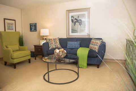 A cozy living room with a blue sofa adorned with patterned pillows and a green throw blanket, a green armchair, a round glass coffee table with decorative items, a wooden side table with a lamp, and framed artwork on the walls.