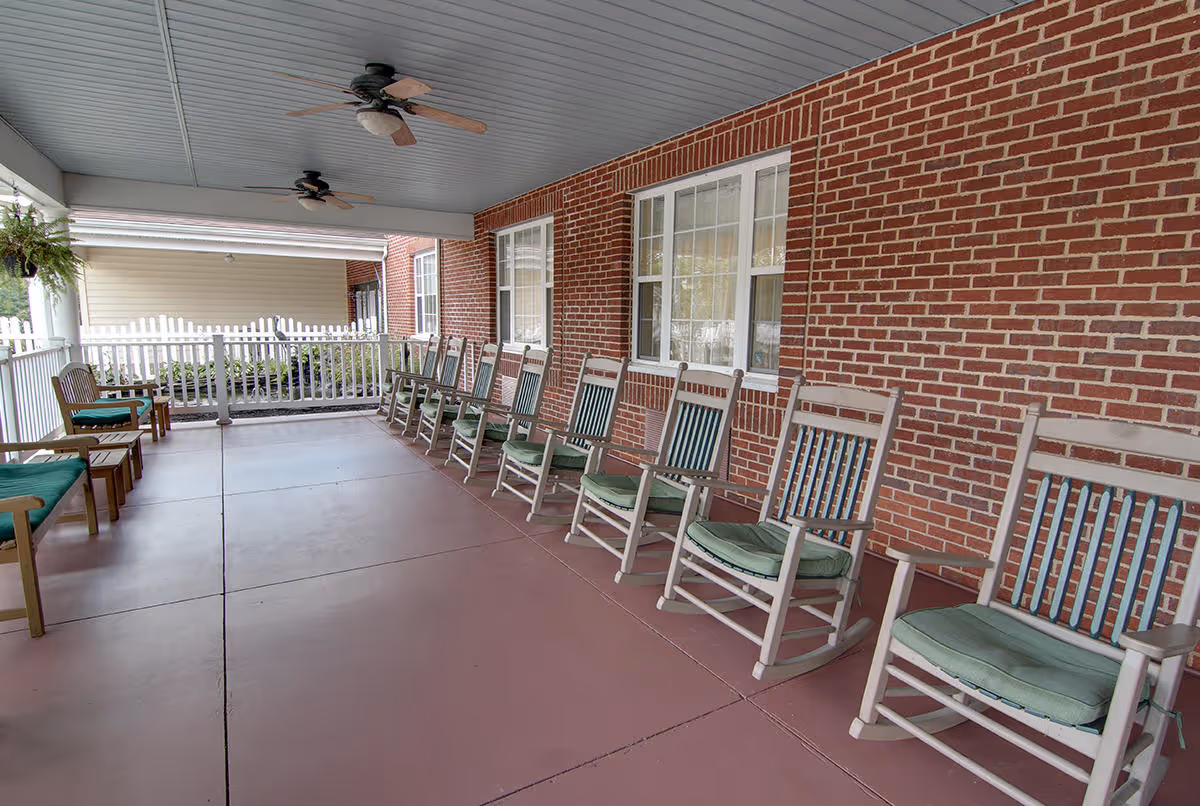 Covered outdoor porch area with a row of wooden rocking chairs with green cushions along a brick wall, ceiling fans overhead, and a white picket fence enclosing the space.