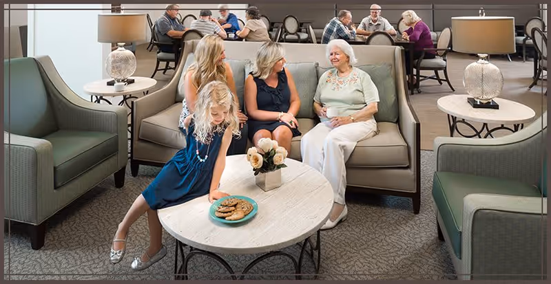 A young girl and three women seated around a coffee table in a bright senior living lounge, with other residents at tables in the background.