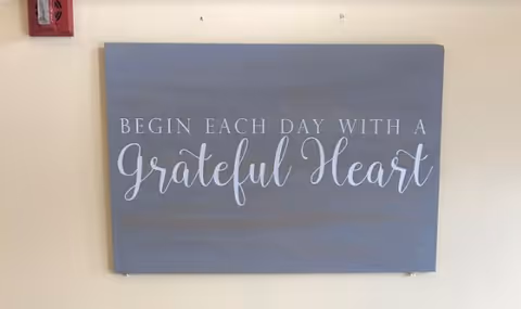 A wall-mounted sign with the inspirational quote 'Begin each day with a grateful heart' written in white cursive and capital letters on a gray background.