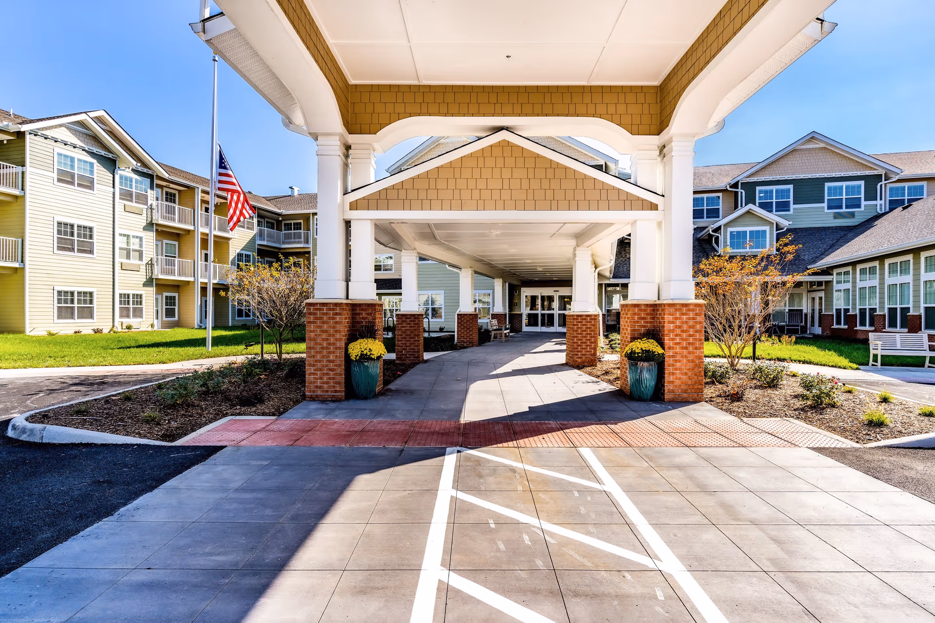 Covered porte-cochere entrance of a multi-story senior living building with landscaping and an American flag.