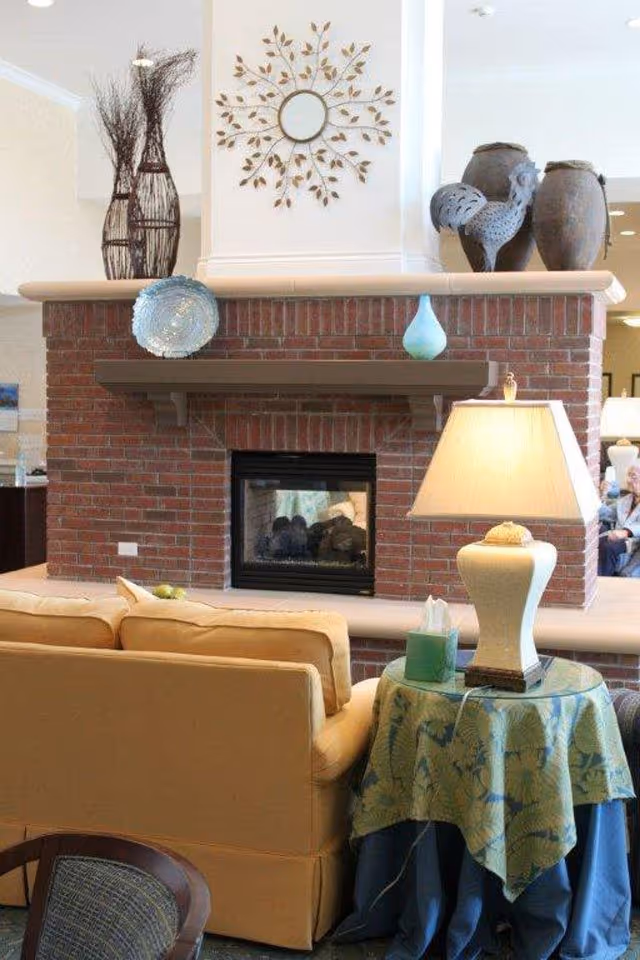 A cozy living room area featuring a brick fireplace with decorative items on the mantel, including a blue plate, a small turquoise vase, and two large pottery pieces shaped like a rooster and a jar. Above the fireplace is a decorative wall mirror with a leaf design. In front of the fireplace is a beige sofa, and next to it is a round side table covered with a green and blue patterned cloth, holding a lamp and a box of tissues.