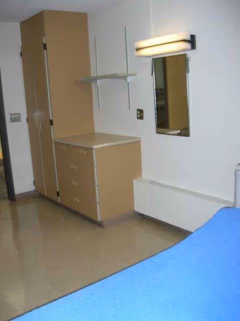 Corner of a bedroom with a beige dresser, wardrobe, mirror, and a blue bed.