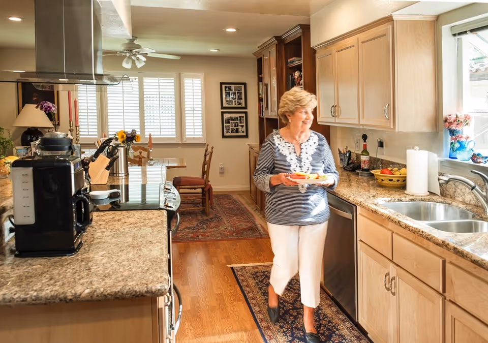 A woman holding a plate of food walking in a bright kitchen with wooden cabinets, granite countertops, a double sink, and various kitchen appliances. The kitchen opens into a dining area with a table, chairs, and windows with white shutters.