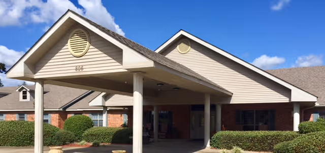 Front entrance of a single-story senior living facility with a covered porte-cochere, brick facade, and landscaped shrubs under a blue sky.