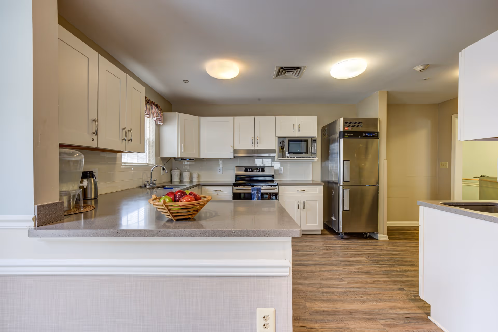 A clean and modern kitchen with white cabinets, stainless steel appliances including a refrigerator, stove, and microwave. The kitchen features a large countertop with a basket of fruit, a water dispenser, and a coffee pot. The floor is wood-style, and the room is well-lit with ceiling lights.