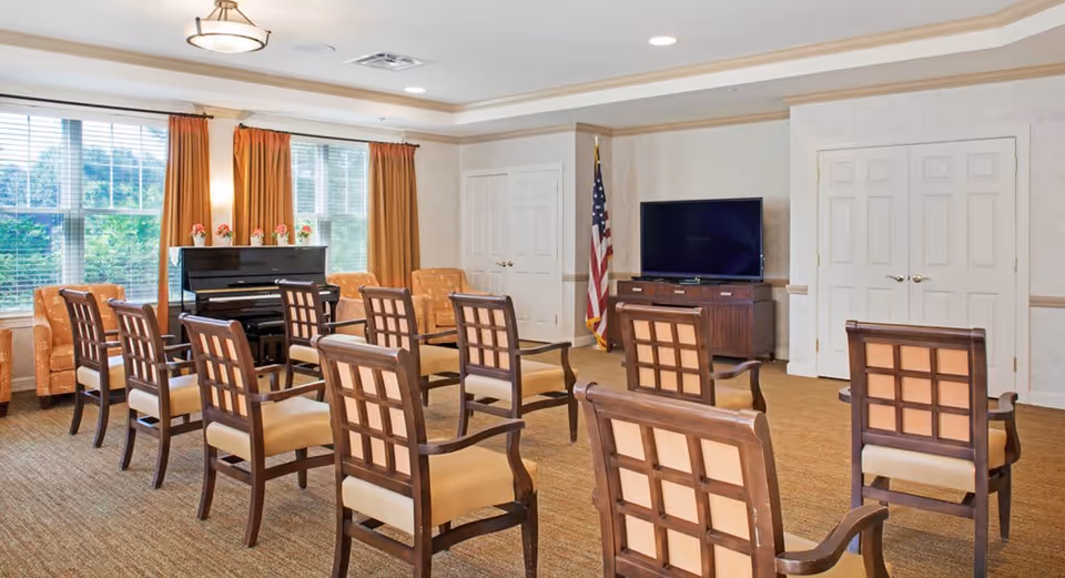 A well-lit common room with multiple wooden chairs arranged in rows facing a flat-screen TV on a wooden cabinet. There is an American flag in the corner, a black upright piano near large windows with orange curtains, and two orange armchairs beside the piano. The room has beige walls and carpeted flooring.
