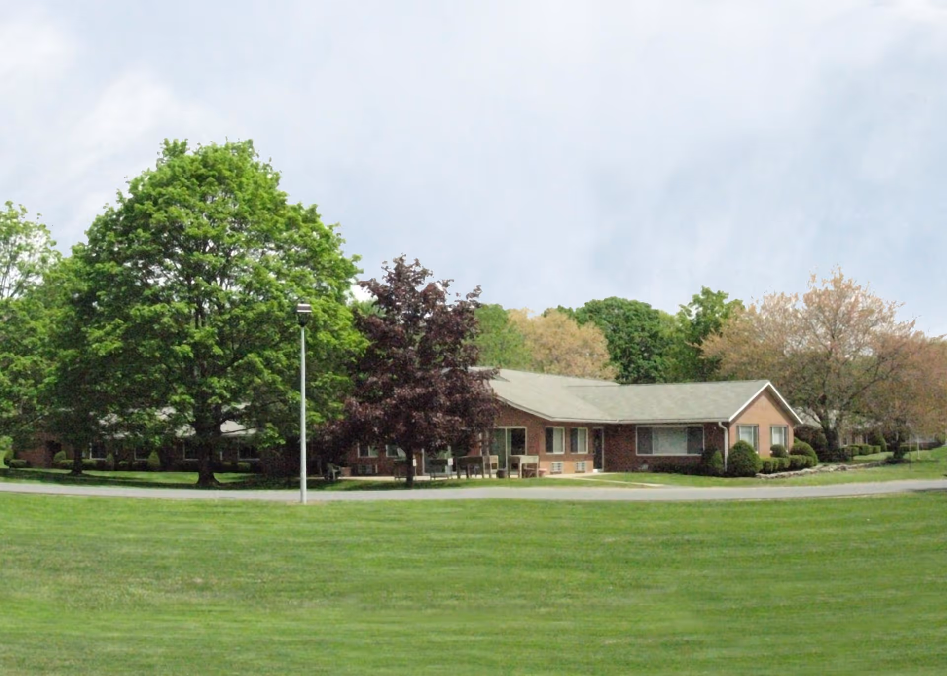 Single-story brick building set behind a wide green lawn with mature trees and a lamppost under a cloudy sky.