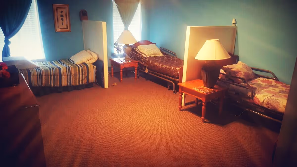 A room with three single beds separated by white partitions. Each bed has different bedding and a pillow. There are two wooden side tables with lamps on them placed between the beds. The room has blue walls, carpeted floor, and two windows with curtains allowing natural light in.