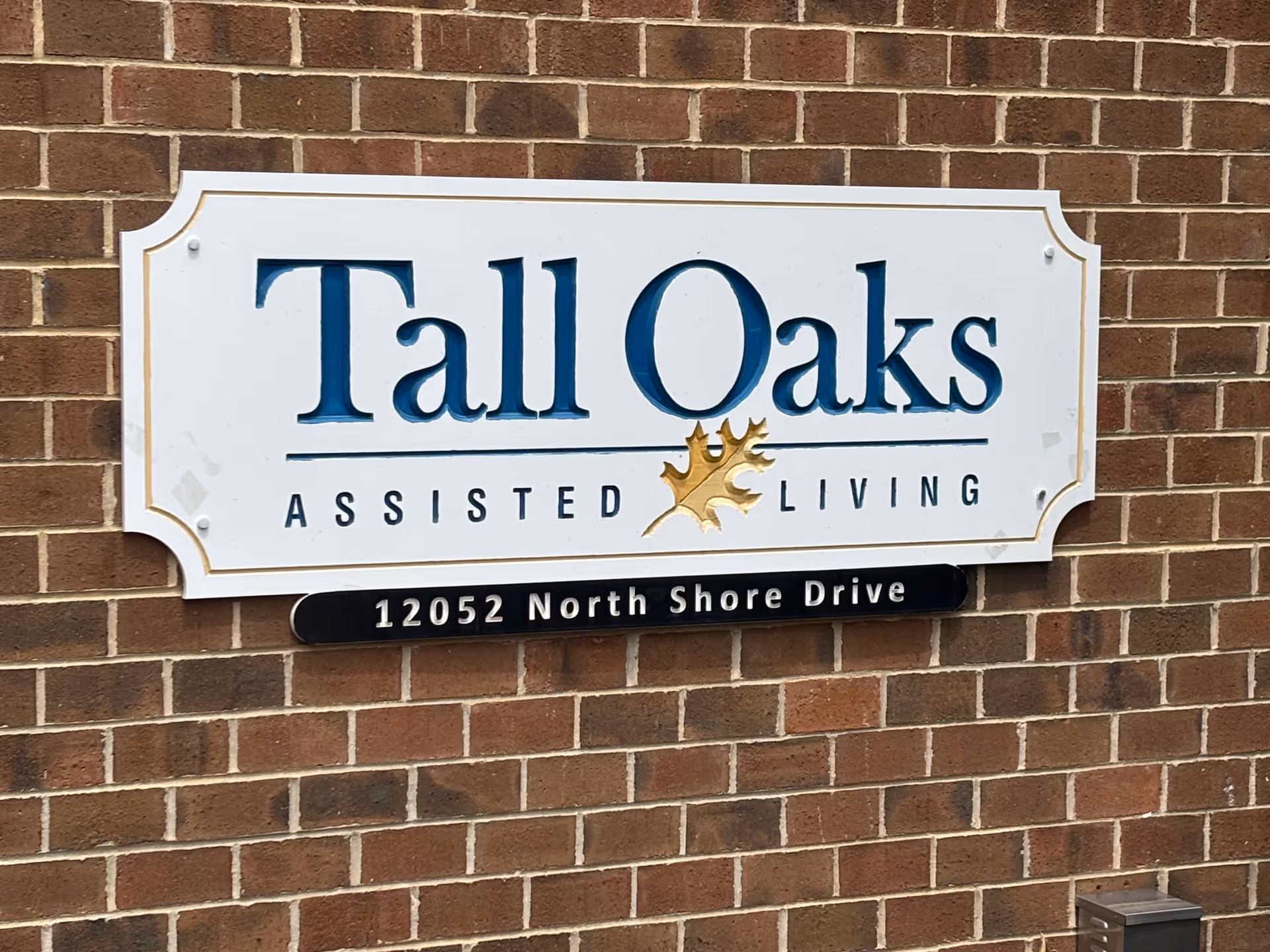 White sign reading "Tall Oaks Assisted Living" with the address "12052 North Shore Drive" mounted on a brick wall.