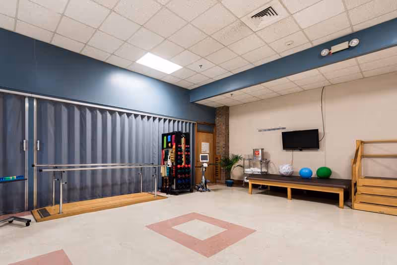Rehabilitation therapy room with parallel bars, exercise equipment, a therapy bench, and a wall-mounted TV.