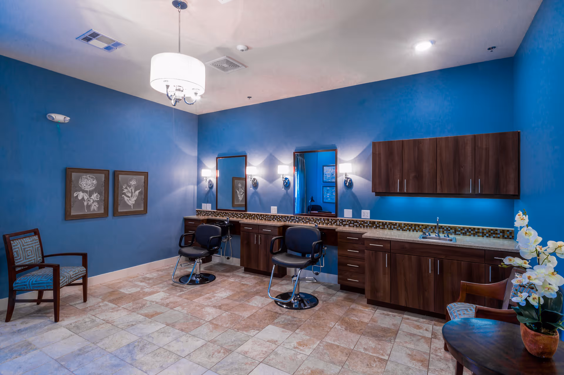 Interior of a salon room with blue walls, two salon chairs in front of mirrors with wall-mounted lights, wooden cabinets, a sink, two framed floral artworks on the wall, and two upholstered chairs with a small round table holding a potted orchid.