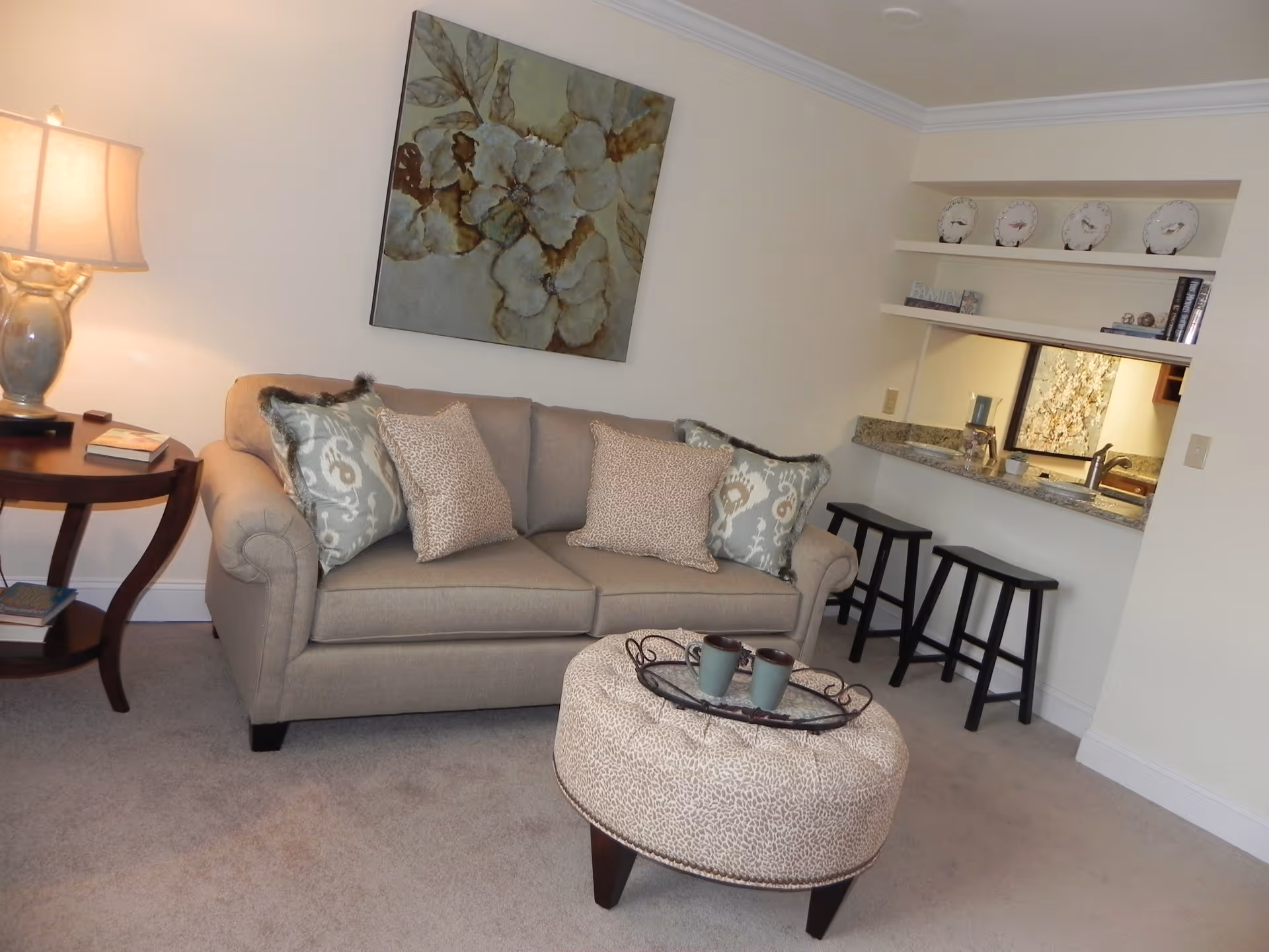 Cozy living room area with a beige sofa adorned with patterned throw pillows, a round ottoman with a tray holding two mugs, a side table with a lamp and books, and a pass-through window to the kitchen with two black bar stools. A large floral painting hangs on the wall above the sofa.