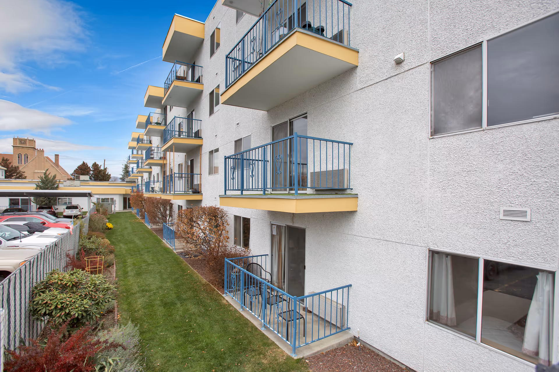 Exterior view of a multi-story senior living facility with balconies on each floor. The building has a white textured facade with blue railings and yellow trim. There is a narrow grassy area with shrubs and a parking lot with cars on the left side. The sky is partly cloudy.
