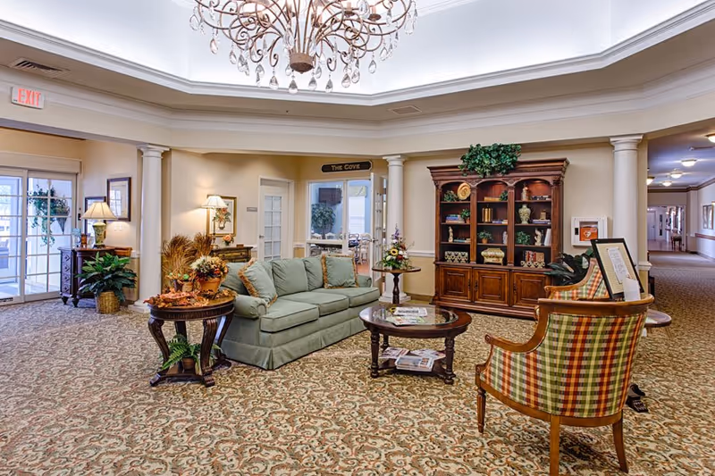 A cozy senior living facility common area with a green sofa, a plaid armchair, two round wooden tables with floral arrangements, a wooden bookshelf filled with decorative items and books, and a large chandelier hanging from a domed ceiling. The room has patterned carpet and cream-colored walls with columns. There is a hallway and glass doors leading to other rooms.