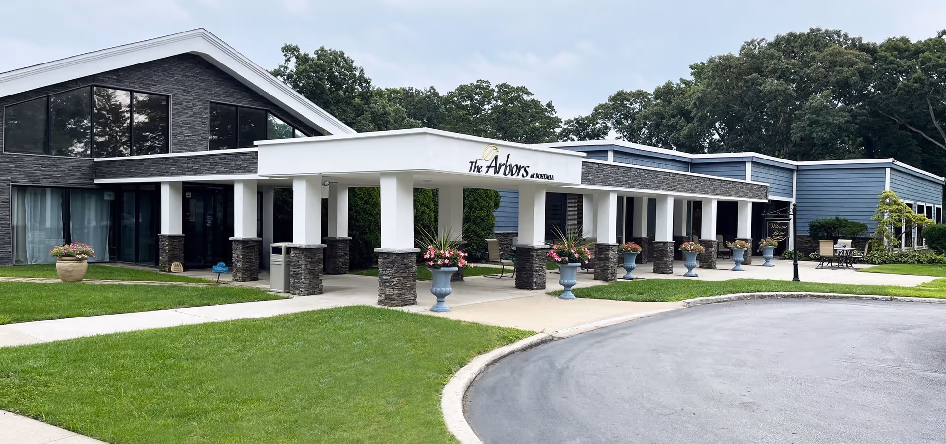 Exterior view of The Arbors senior living facility showing a modern building with a covered entrance supported by white columns with stone bases. There are flower pots with pink flowers along the walkway, green grass, and trees in the background.
