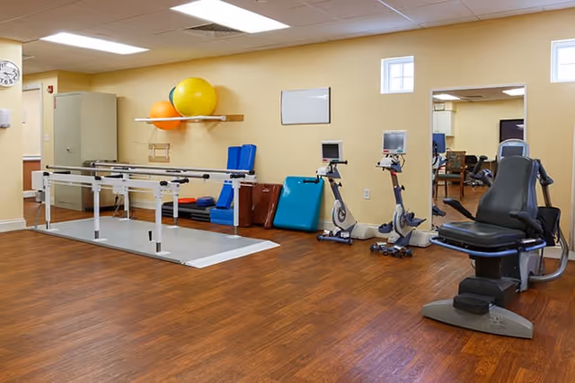 A physical therapy or exercise room with wooden flooring, parallel bars for walking exercises, two stationary exercise bikes, a recumbent exercise machine, and various exercise mats and large colorful exercise balls on a wall shelf. A large mirror is mounted on the wall reflecting part of the room.
