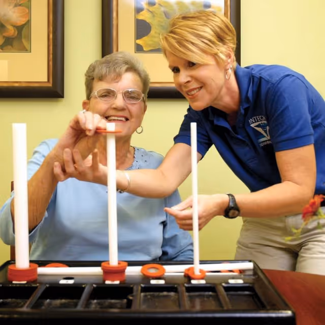 An elderly woman and a caregiver playing a ring toss game together indoors. The elderly woman is smiling and placing a ring on a peg while the caregiver, wearing a blue shirt, assists and smiles.