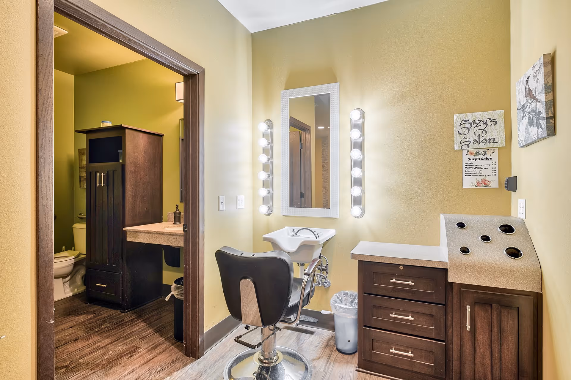 A small salon room with a styling chair in front of a mirror framed by two vertical rows of light bulbs. To the right is a countertop with drawers and a slanted section with holes for holding salon tools. A sign on the wall reads 'Suzy's Salon'. Through an open doorway on the left, a bathroom with a toilet and a dark wooden cabinet is visible.