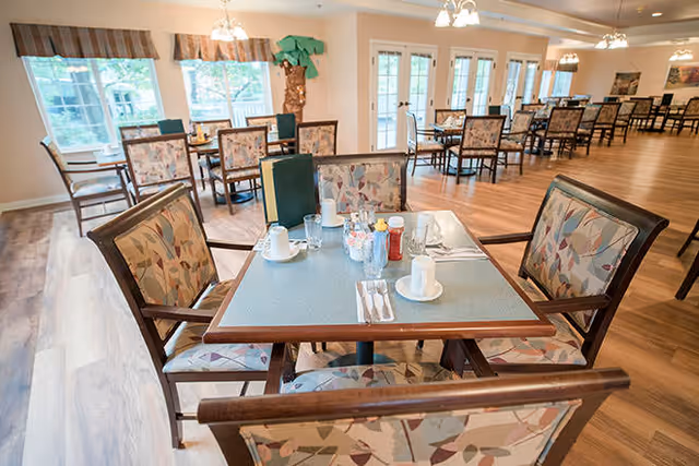 Bright dining room with several tables and patterned chairs set for a meal in a communal senior living space.