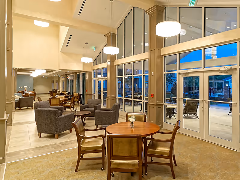 A spacious and well-lit common area in a senior living facility featuring multiple seating arrangements including round wooden tables with chairs and cushioned armchairs. Large floor-to-ceiling windows and glass doors provide a view of an outdoor patio area with additional seating. The room has high ceilings with modern hanging light fixtures and neutral-toned walls and flooring.