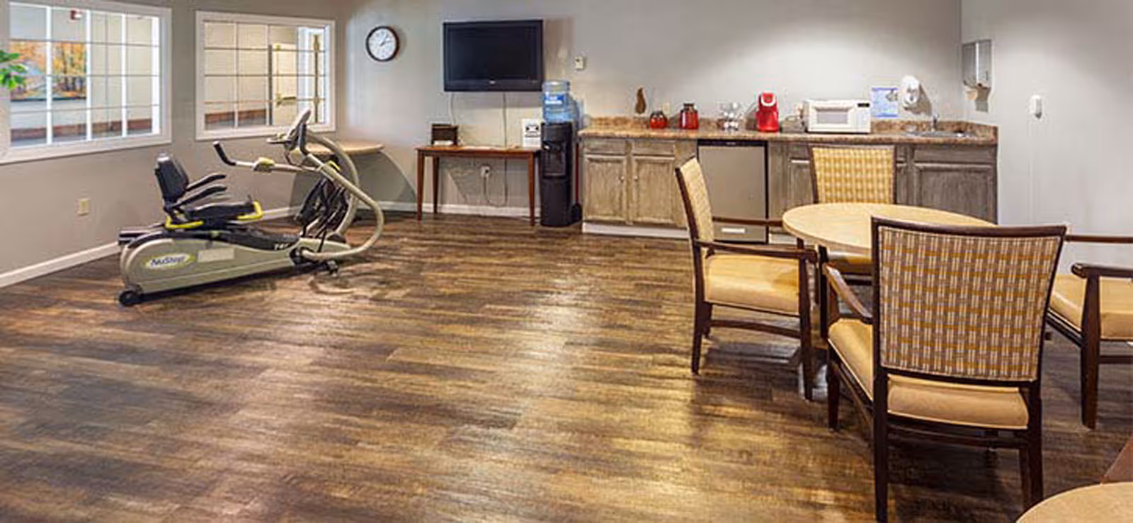 Common interior community room with a kitchenette, round table and chairs, wall-mounted TV, water cooler and exercise equipment on wood-look flooring.