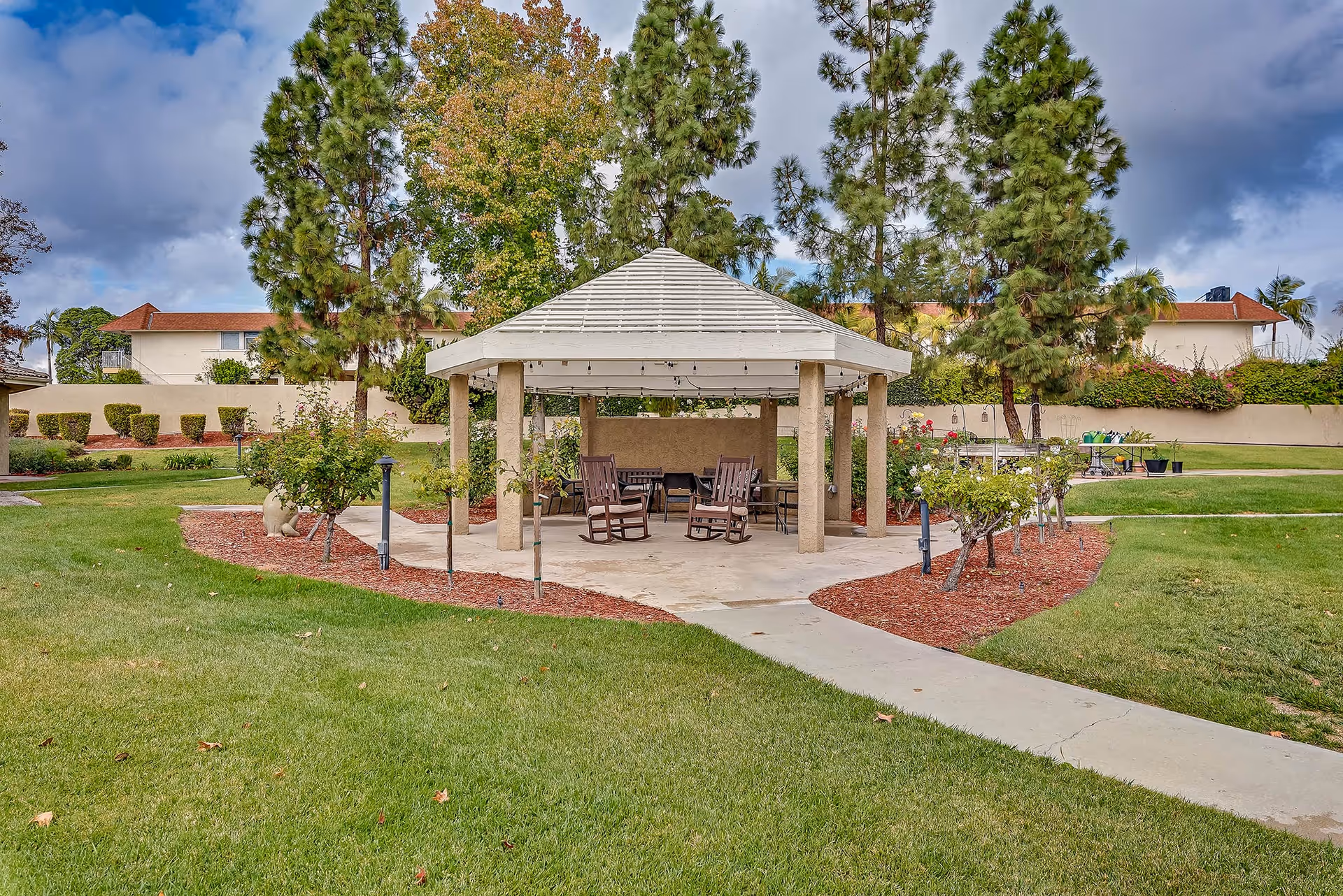 Outdoor gazebo structure with a white roof and beige pillars surrounded by green grass, small trees, and shrubs. There are several wooden rocking chairs and tables under the gazebo. Residential buildings and tall pine trees are visible in the background under a partly cloudy sky.