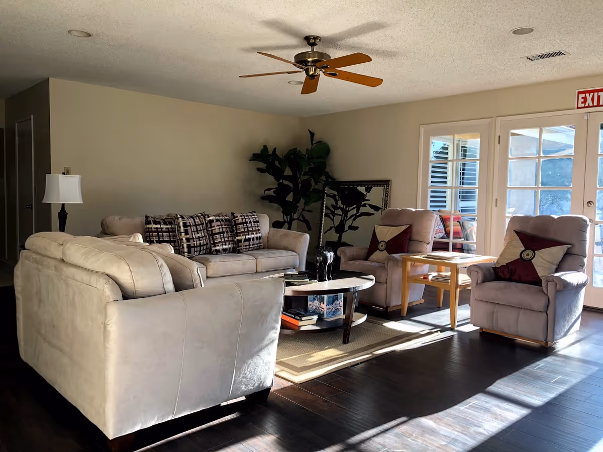 Sunlit living room with beige sofas and armchairs arranged around a round coffee table near glass doors and a ceiling fan.