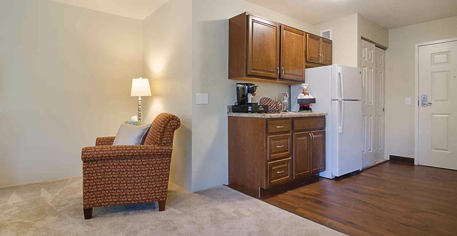 A cozy corner of a senior living facility apartment featuring a patterned armchair with a pillow and a floor lamp beside it. Adjacent to the seating area is a small kitchen space with wooden cabinets, a countertop holding a coffee maker and decorative items, and a white refrigerator. The floor transitions from carpet in the seating area to wood in the kitchen area. A white door and closet with sliding doors are visible in the background.