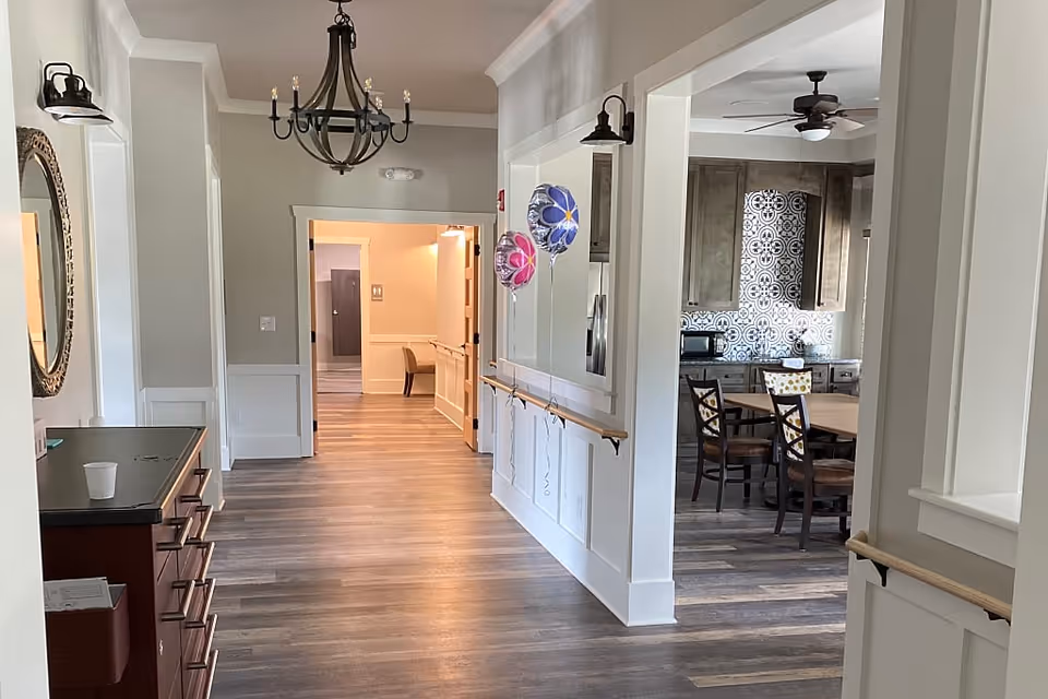 Interior view of a senior living facility hallway with wooden flooring and white walls. On the right side, there is an open kitchen area with a dining table and chairs, featuring a decorative tiled backsplash and ceiling fan. The hallway has a chandelier light fixture and handrails along the walls. Two colorful balloons are tied to the handrail on the right side. A round mirror and a small cabinet with drawers are visible on the left side of the hallway.