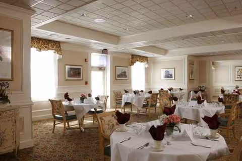 Elegant senior living dining room with round tables, white linens, burgundy napkins, and framed wall art.