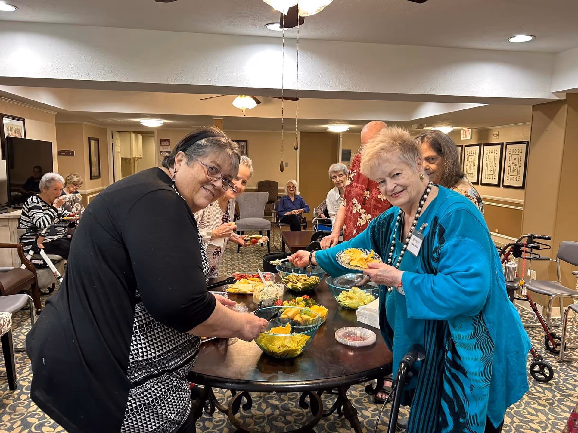A group of elderly people gathered around a table with bowls of snacks and fruit in a common area of a senior living facility. Some are serving themselves food while others are seated in the background. The room has warm lighting and comfortable seating.