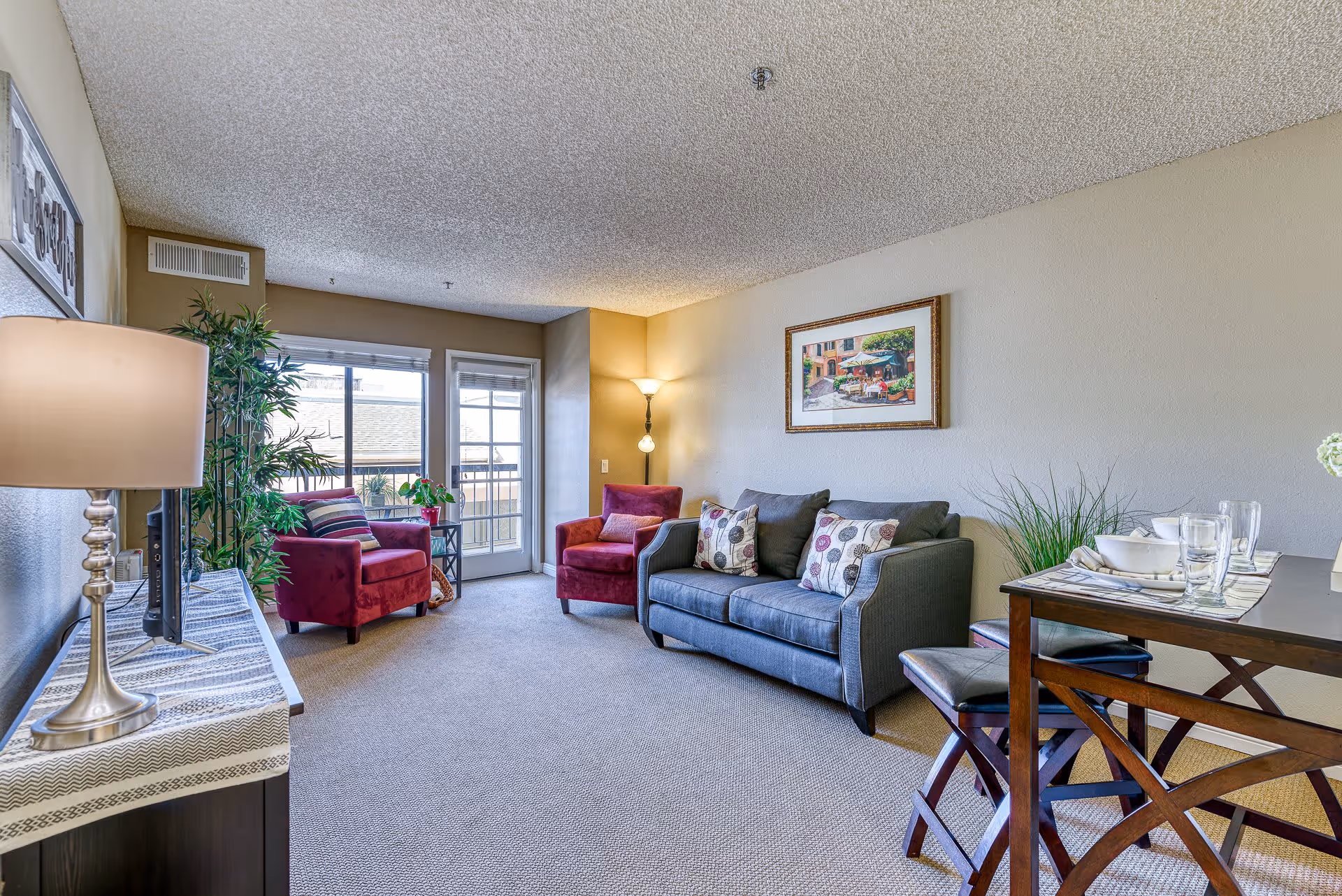 A cozy senior living room featuring a gray sofa with patterned cushions, two red armchairs, a floor lamp in the corner, a framed painting on the wall, a dining table set with dishes and glasses, a TV on a stand with a table lamp, and a large window with a glass door leading outside.