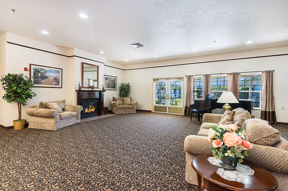 A spacious living room in a retirement community featuring beige sofas, a patterned carpet, a fireplace with a mirror above it, framed artwork on the walls, potted plants, a grand piano near large windows with curtains, and a wooden side table with a floral arrangement and a lamp.