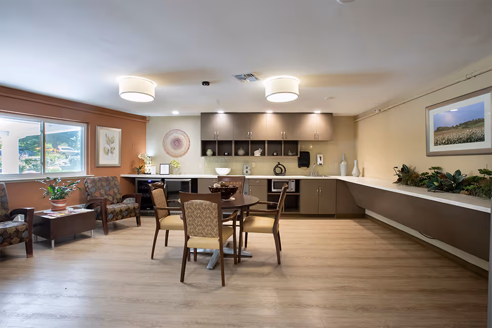 A well-lit common area with a round table and four chairs in the center, a kitchenette with cabinets, a microwave, and decorative items along the back wall. To the left, there are two patterned armchairs with a small table between them holding a potted plant. A large window lets in natural light, and the walls are decorated with framed artwork and plants.