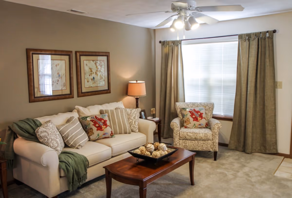 A cozy living room with a beige sofa adorned with multiple decorative pillows and a green throw blanket. Next to the sofa is a patterned armchair with a colorful pillow. A wooden coffee table with decorative spheres sits in front of the sofa. The room has beige walls with two framed pictures, a window with closed blinds and green curtains, a ceiling fan with lights, and a table lamp on a side table.