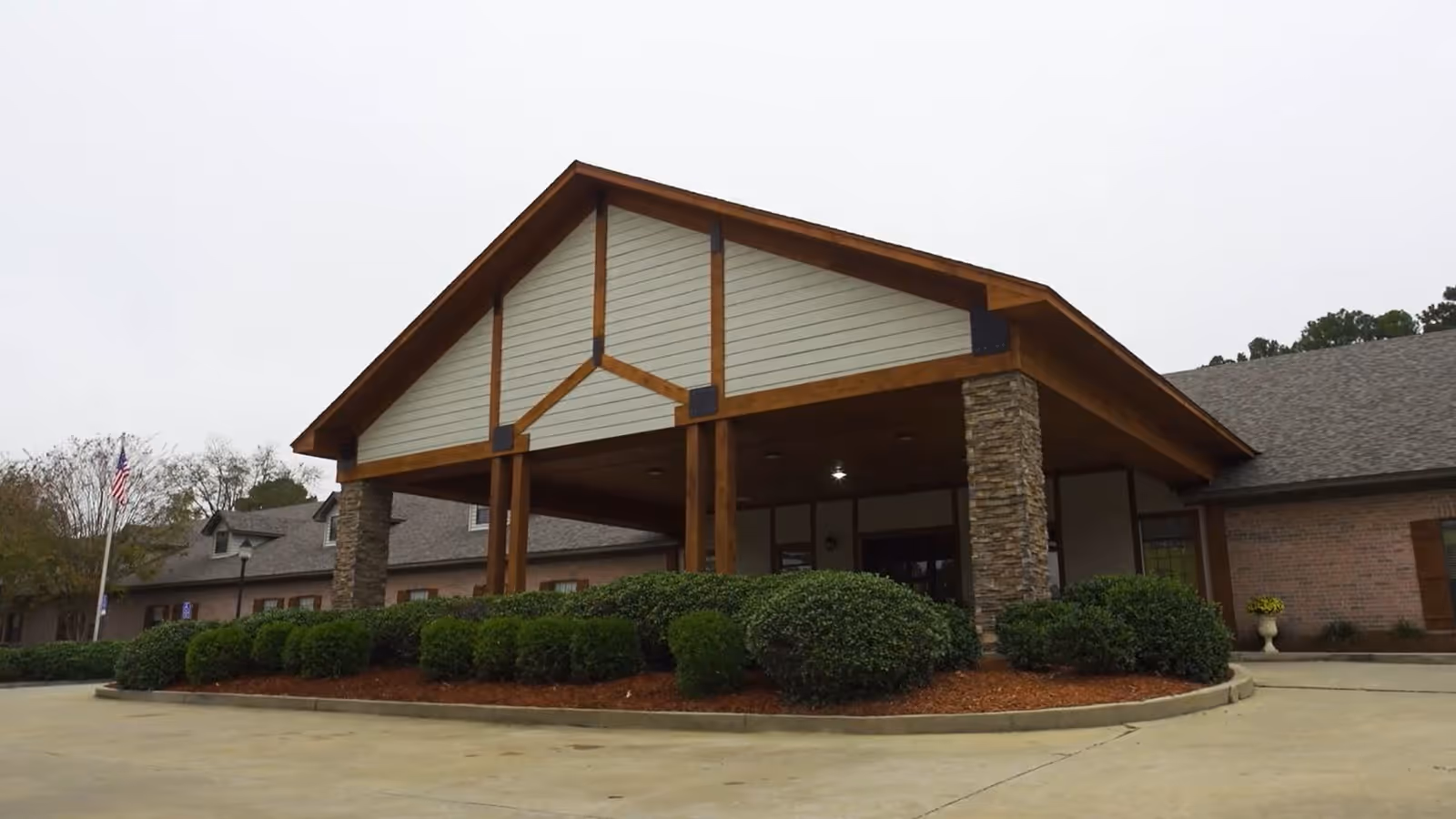 Exterior front view of Arbor Lake Skilled Nursing and Rehabilitation Center showing a covered entrance with wooden beams and stone pillars, surrounded by neatly trimmed bushes and a concrete driveway.