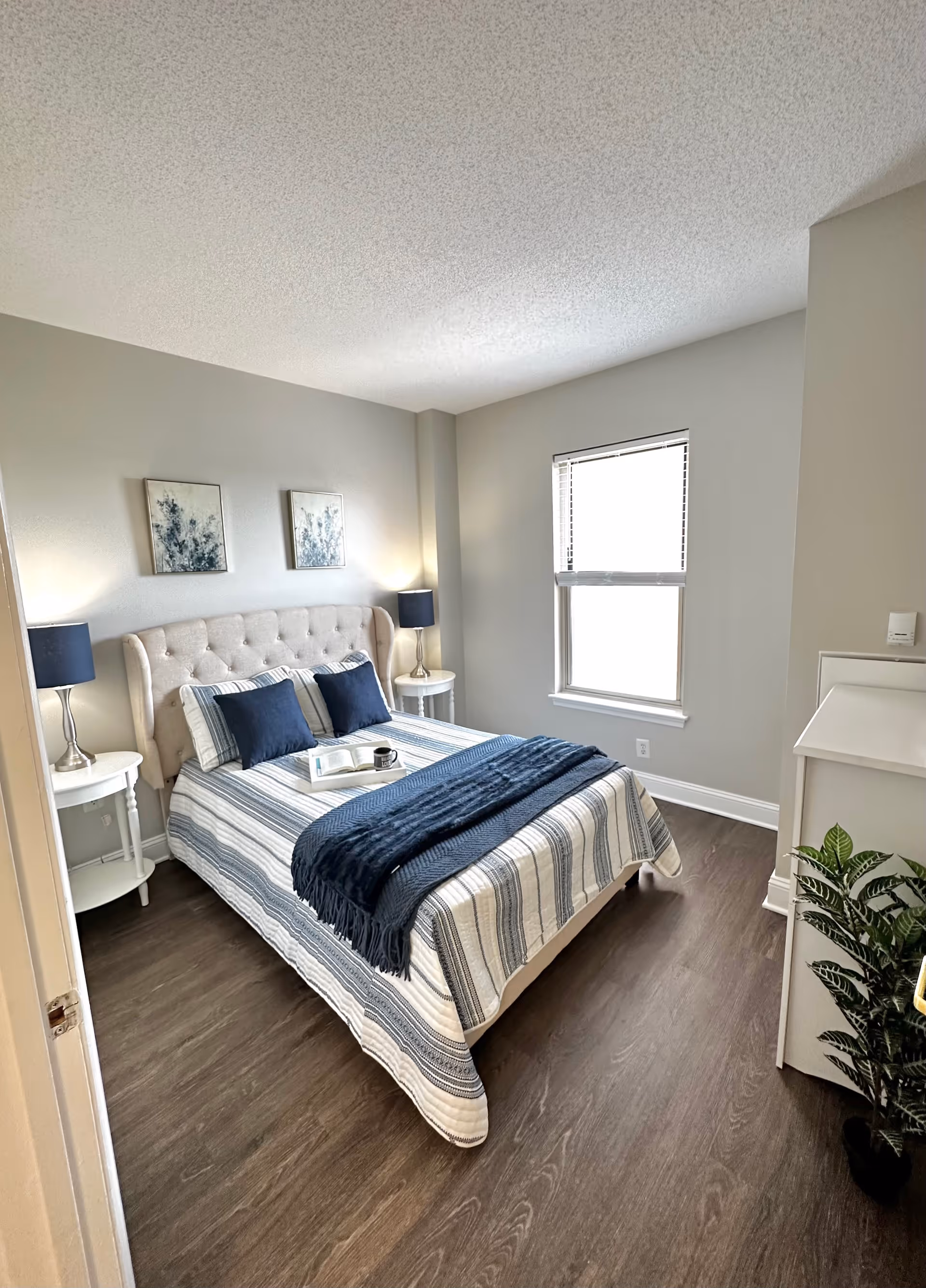 A bright bedroom with a bed featuring a tufted beige headboard, striped bedding in shades of blue and white, and two navy blue pillows. There are two matching bedside tables with silver lamps and dark blue lampshades. Two framed abstract artworks hang above the bed. A window with blinds lets in natural light, and a small white cabinet with a green plant on the floor is visible in the corner. The room has light gray walls and dark wood flooring.