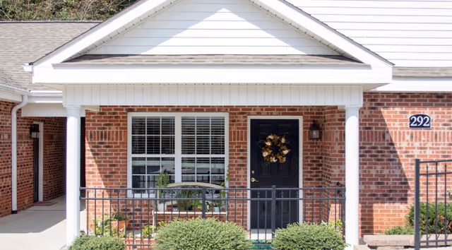 Front exterior view of a brick building with a white porch supported by two white columns. There is a black door with a decorative wreath and a window with white blinds next to it. The building number 292 is visible on the right side of the door. Green shrubs are planted in front of the porch, and a black metal railing encloses the porch area.