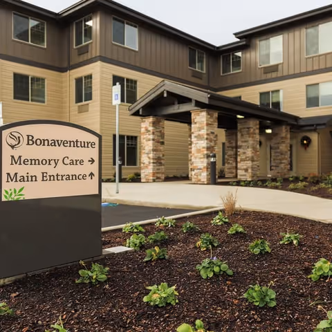 Exterior view of Bonaventure senior living facility showing the main entrance with a covered drop-off area supported by stone pillars. In the foreground, there is a landscaped area with small plants and a sign indicating directions to Memory Care and Main Entrance.