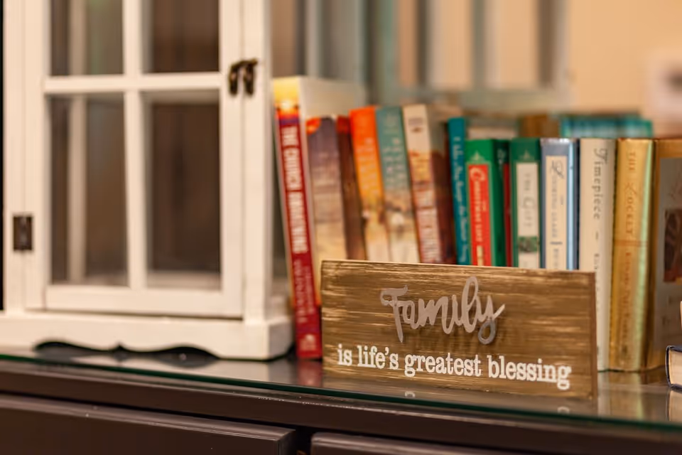 A close-up view of a shelf with a small white decorative cabinet, several books standing upright, and a wooden sign that reads 'Family is life's greatest blessing.'