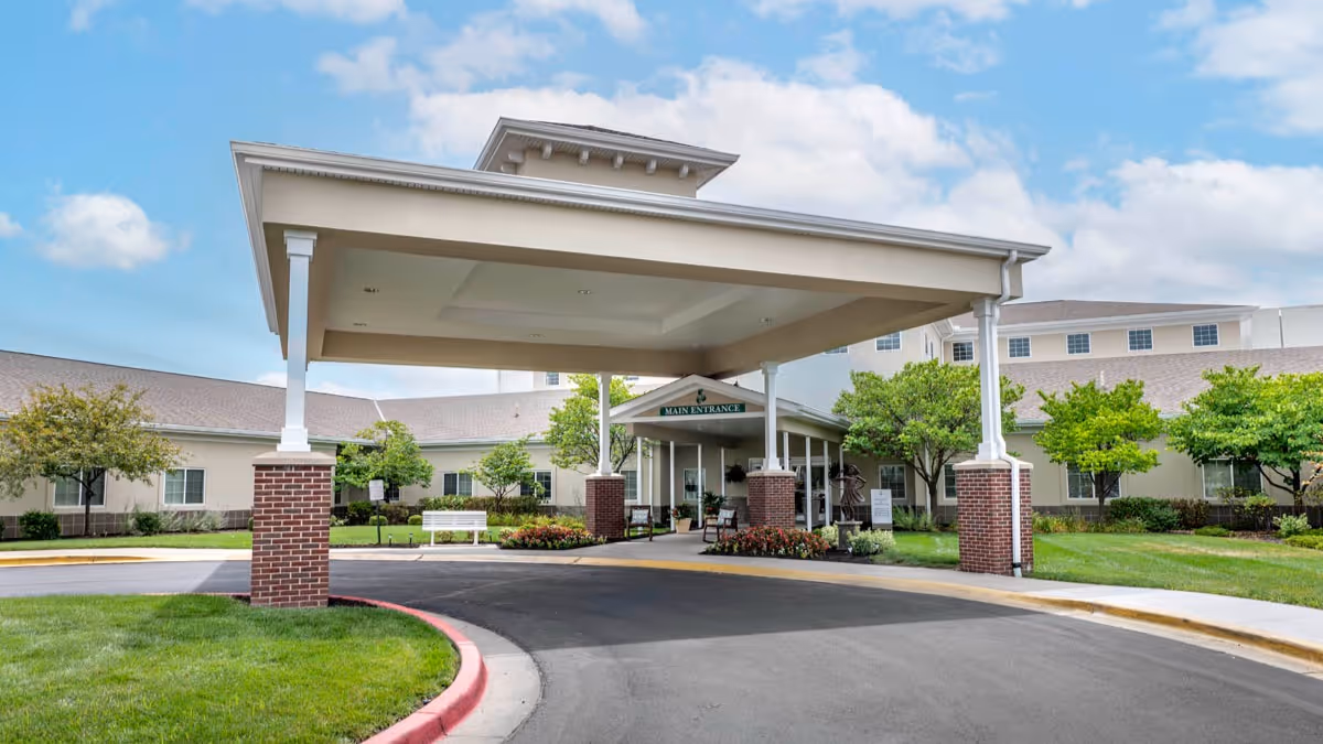 Front exterior view of Brookdale Overland Park senior living facility showing a covered entrance with brick pillars, a driveway, green lawns, trees, and a partly cloudy sky.