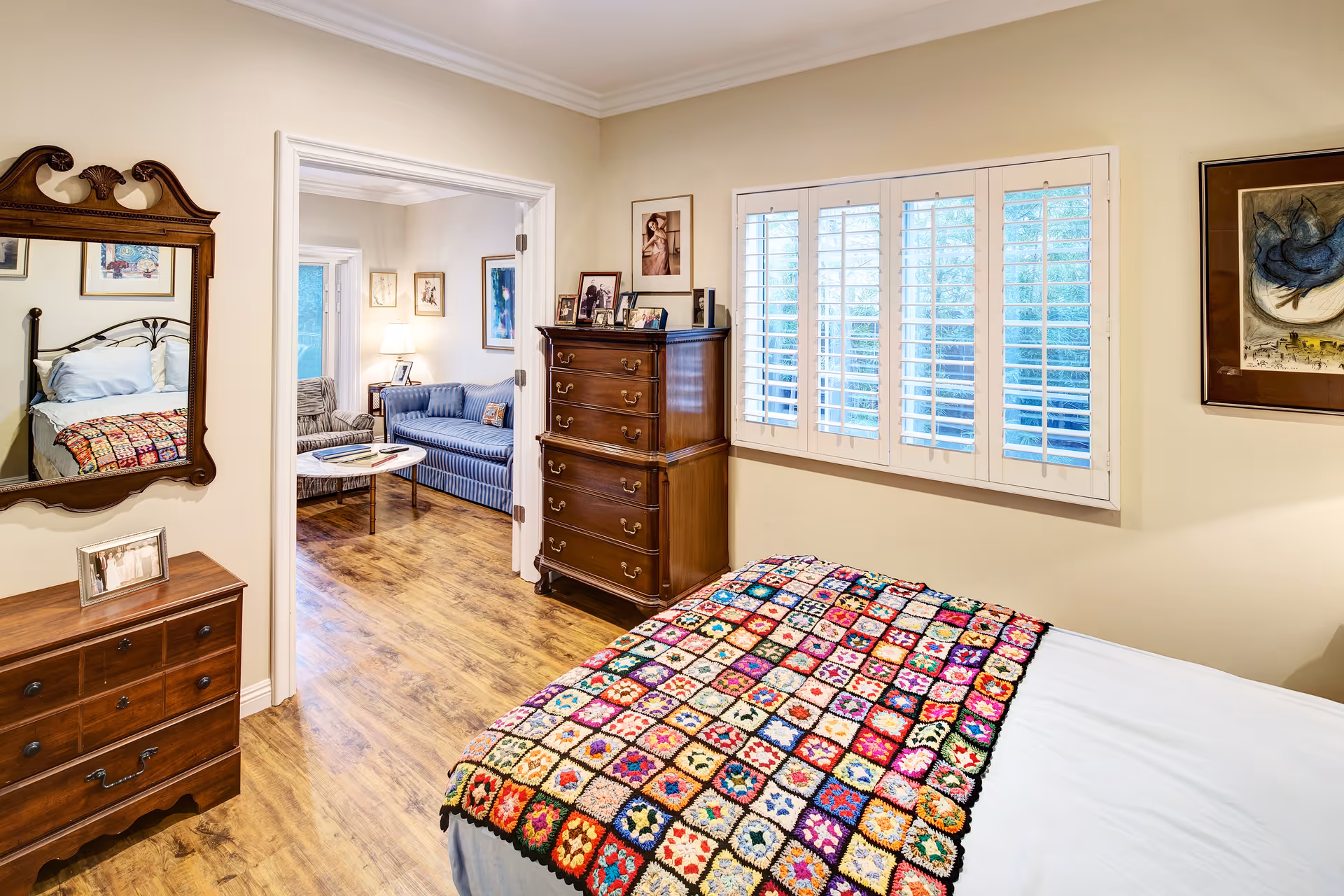 A cozy bedroom with a bed covered by a colorful crocheted blanket. The room has wooden dressers, framed pictures on the walls, and a window with white shutters. Through an open doorway, a living room with a blue striped sofa, an armchair, a coffee table, and framed artwork is visible. The floor is wooden throughout.