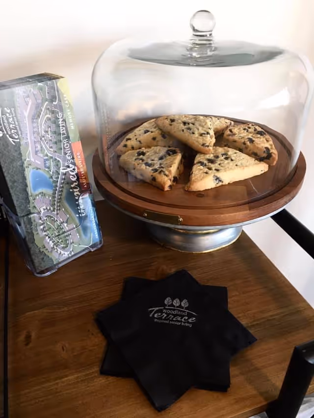 A wooden table with a glass dome covering six triangular chocolate chip scones on a wooden tray. Next to the tray is a brochure for Woodland Terrace senior living and two black napkins with the Woodland Terrace logo and the text 'Inspired senior living'.
