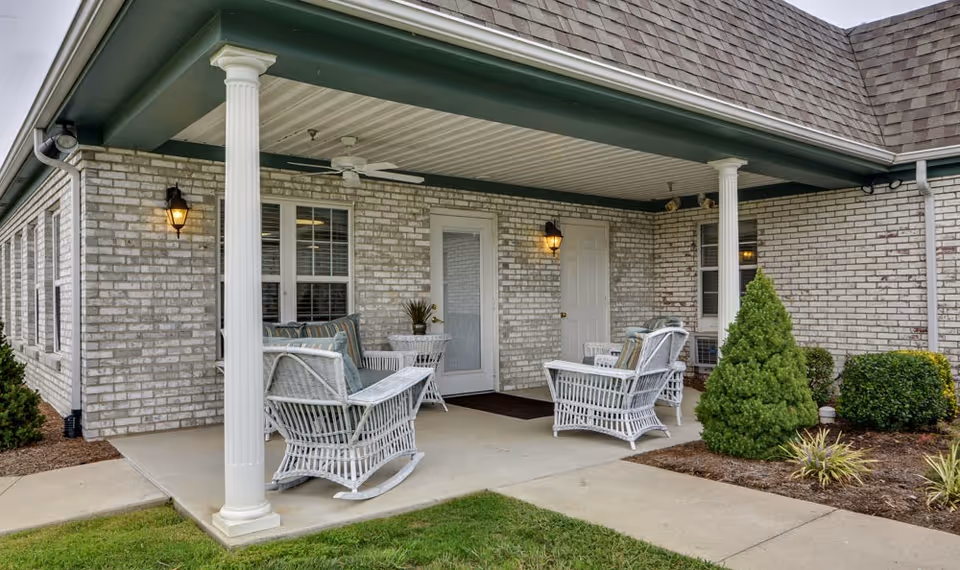 Covered outdoor patio area at a senior living facility with white wicker rocking chairs and a small table. The patio has white columns, ceiling fans, and wall-mounted lantern lights. Surrounding the patio are green shrubs and a well-maintained lawn.